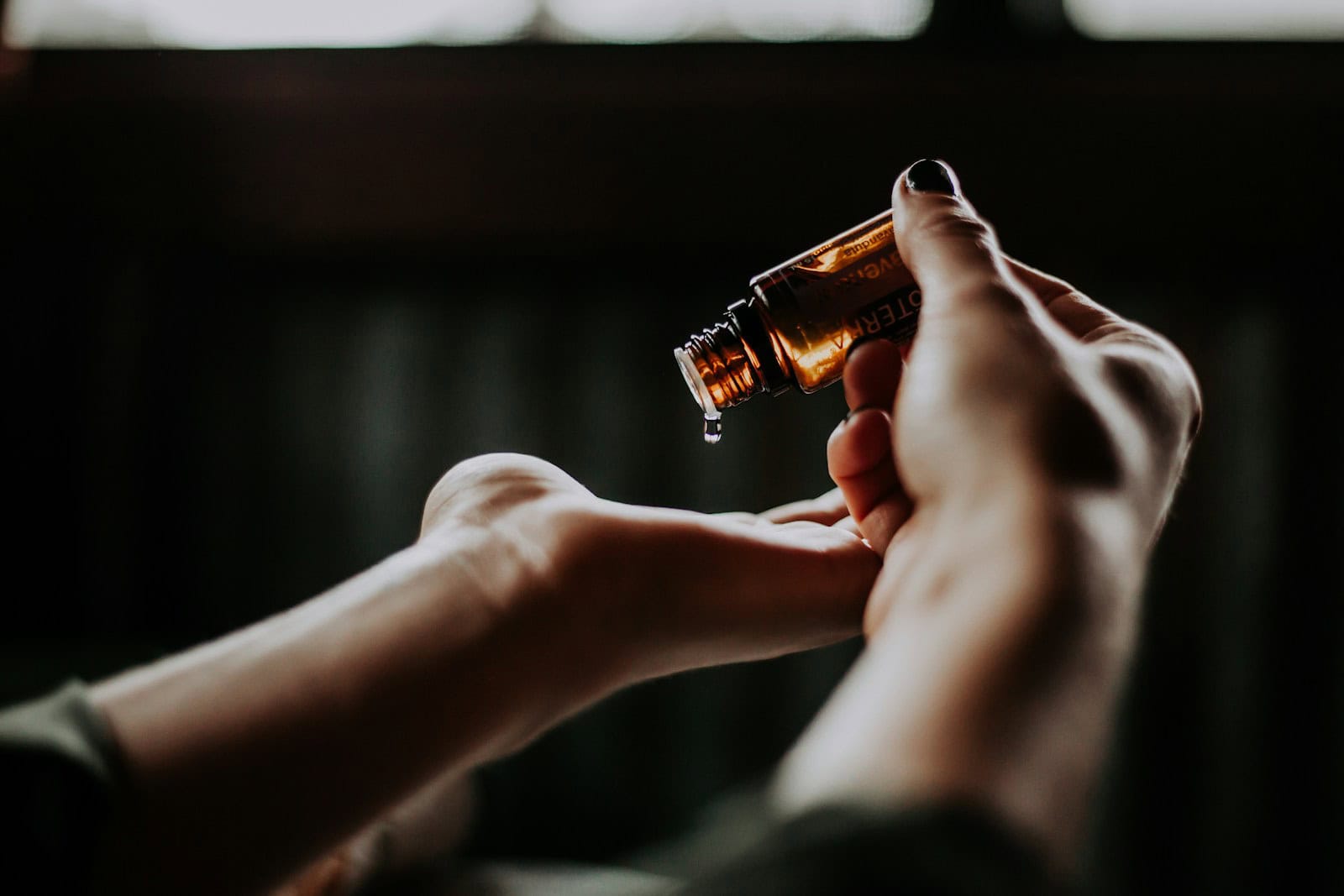 Contenant d'huile essentielle en verre brun avec gouttelette, mains de femme, huile pour bien-être, aromathérapie, relaxation, produit naturel, santé holistique, soins du corps, bien-être à domicile.