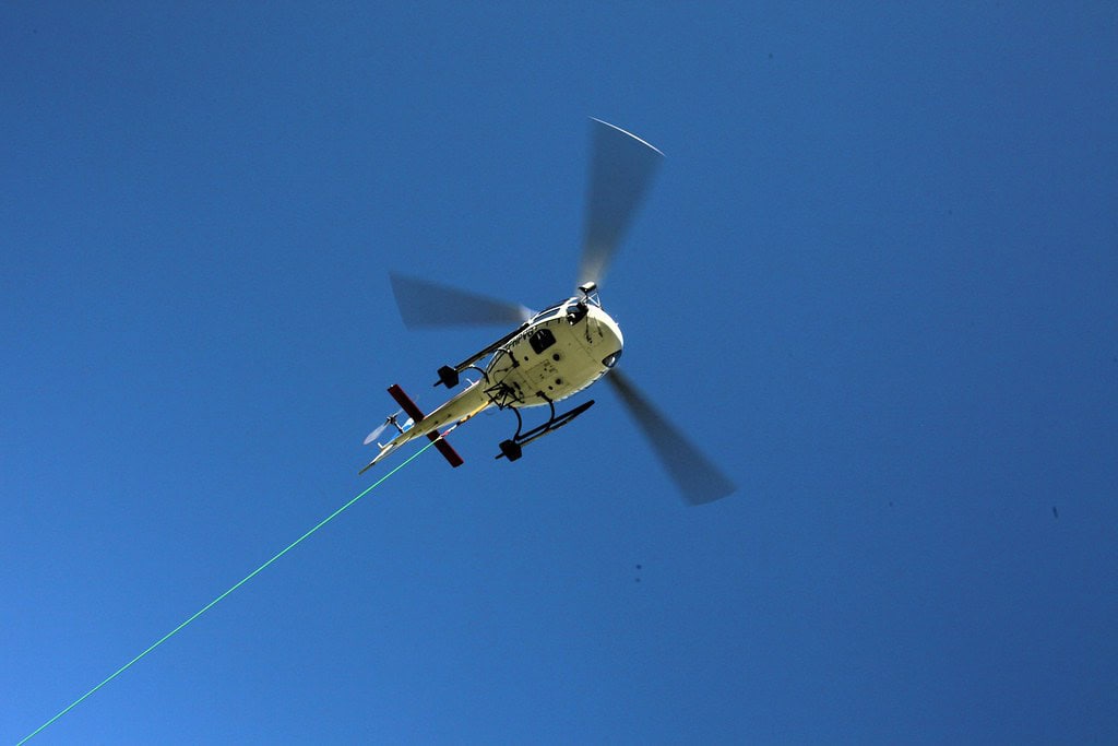 Hélicoptère en vol avec câble de levage contre un ciel bleu clair, symbolisant la sécurité et l'intervention aérienne.