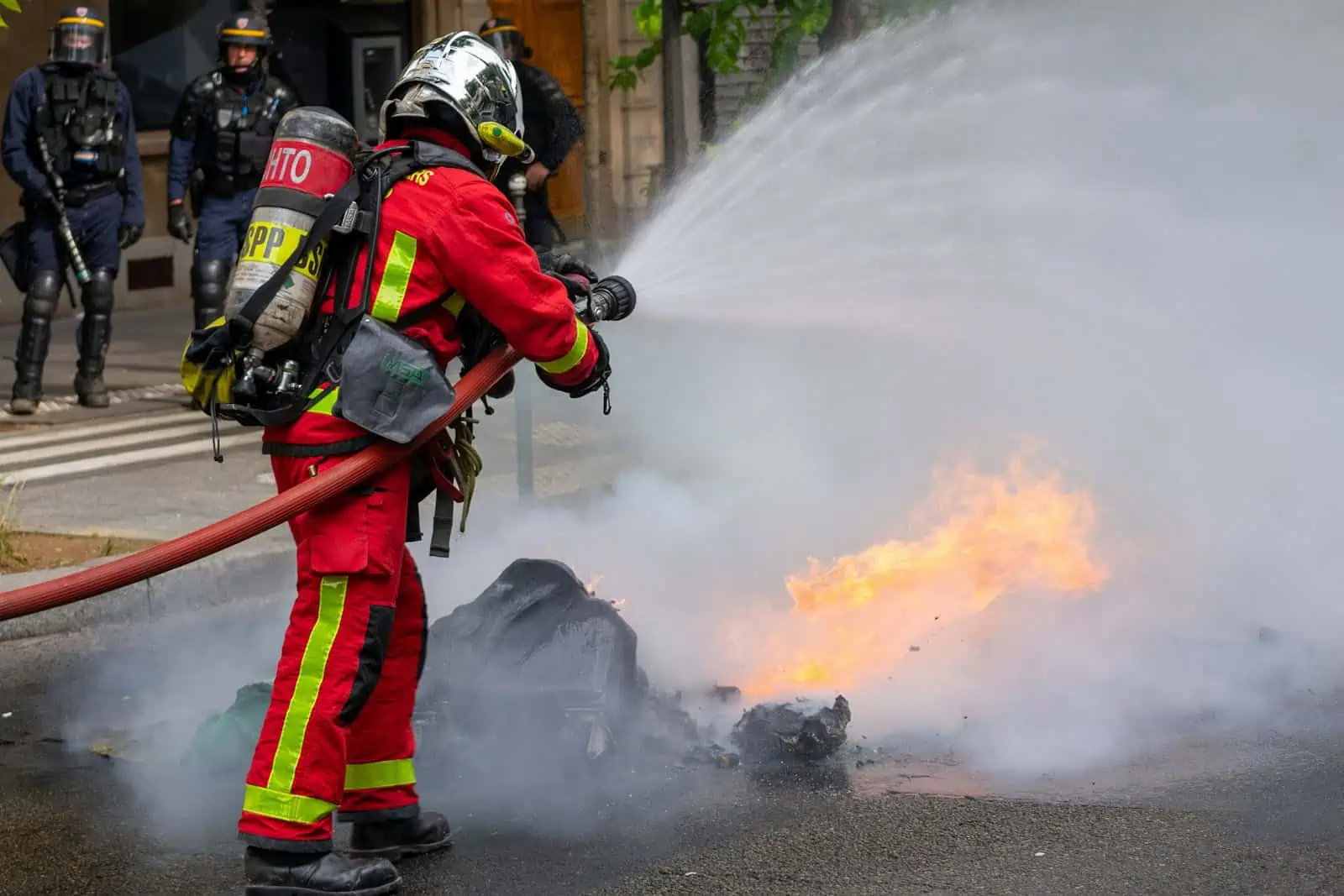 Extérieur, pompier en action éteignant un feu de véhicule avec une lance à haute pression, flames et fumée visibles, sécurité assurée par un équipement spécialisé, intervention d’urgence essentielle.