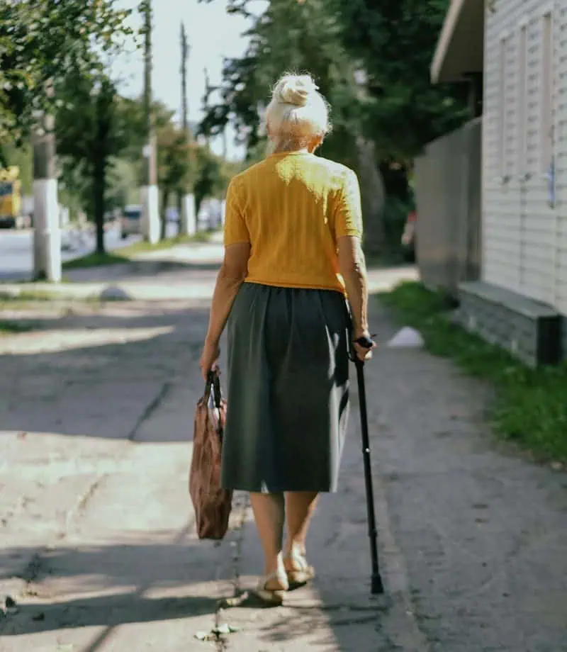 Femme âgée marchant avec une canne et un sac sur un trottoir en ville.