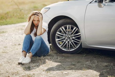 Fille assise au sol avec tête dans les mains à côté d'une voiture arrêtée sur une route poussiéreuse, illustrant l'importance de l'assurance auto à Neuchâtel.